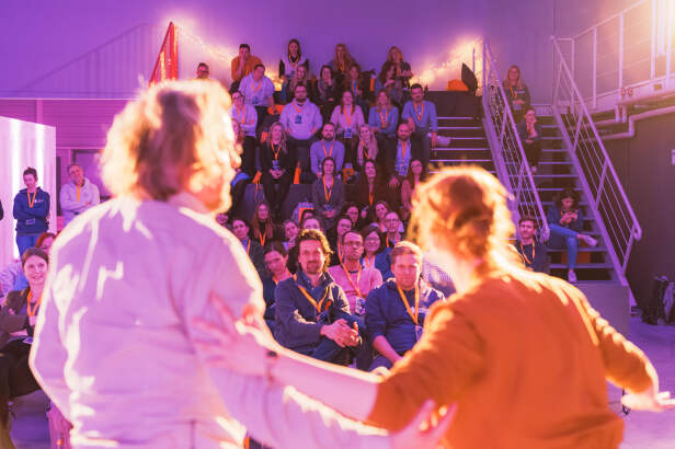 Menschen sitzen im Publikum. Vorne zwei Personen, die unscharf dargestellt sind und auf der Bühne interagieren.